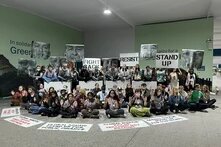 Foto: Eine Gruppe Aktivist*innen sitzt und steht in einer Halle, hält Schilder mit Aufschriften wie „FIGHT BACK“ und „RESIST“ und hebt die Hände.