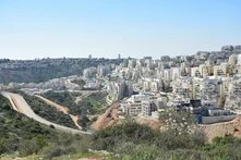Photo: A cityscape with multiple residential buildings along a hill and a road. Green hills and trees in the background. 